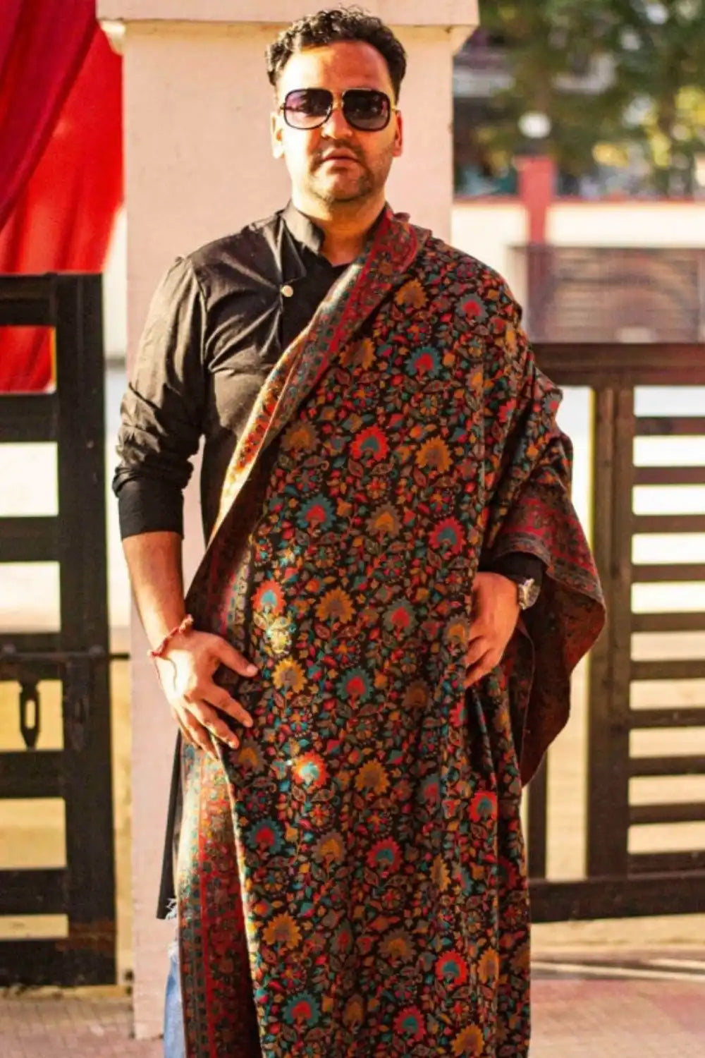 A man wearing a black kurta, sunglasses, and a colorful, intricately patterned shawl.