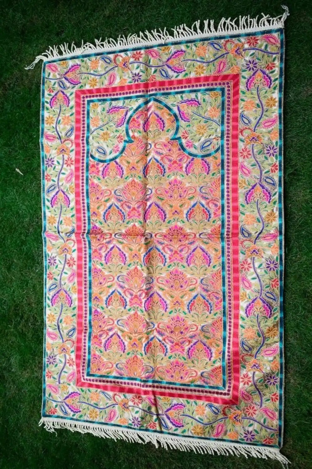 A rectangular prayer rug with intricate floral and paisley patterns in shades of pink, orange, teal, and purple, featuring a central mihrab design and fringed ends.